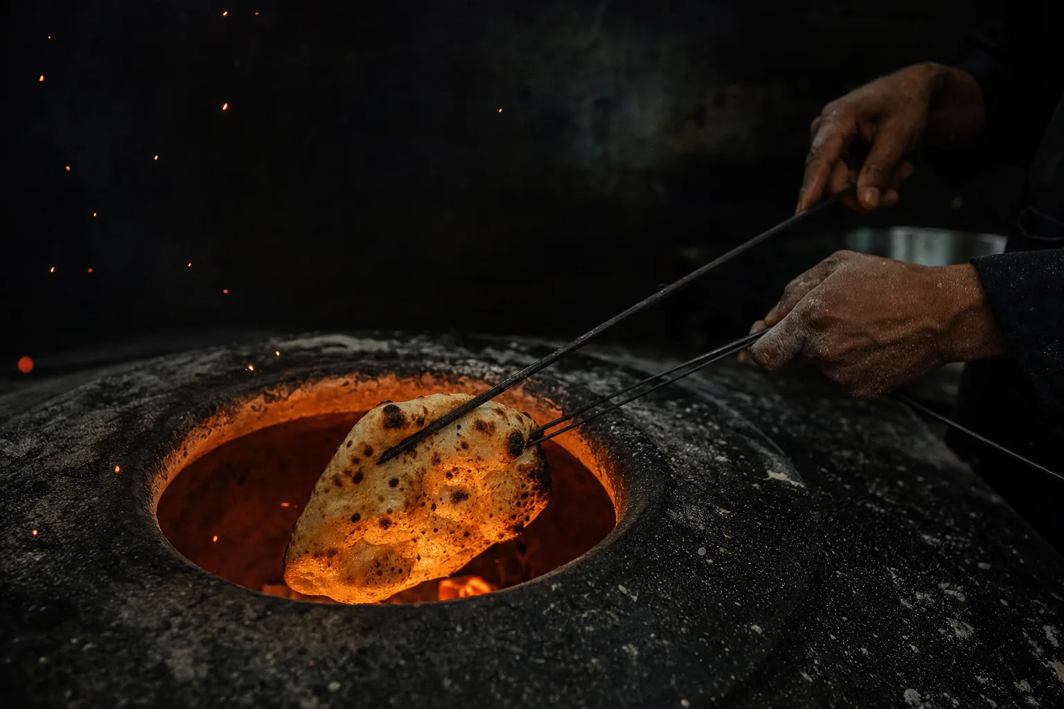 Un cuisinier sort un naan boursouflé d'un tandoor incandescent, dans une cuisine sombre.